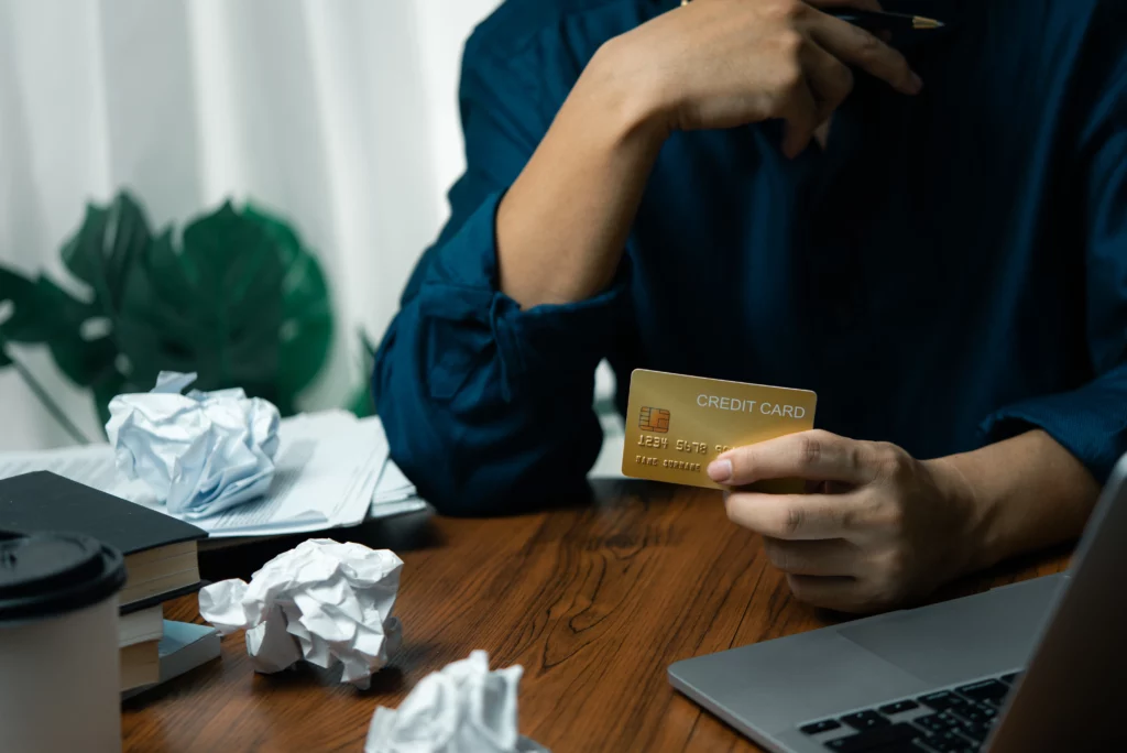 A person holding a gold credit card at a wooden desk with crumpled papers, books, and a laptop, symbolizing financial stress or planning.