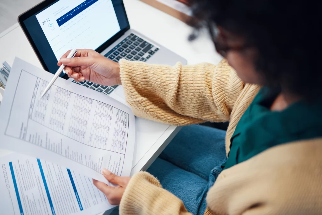 Woman in a yellow sweater reviewing financial documents at a desk with a laptop displaying graphs, focusing on financial recovery.