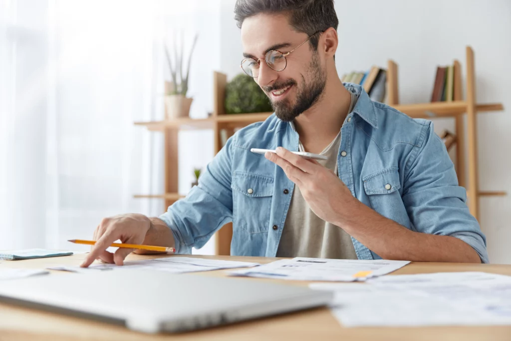 Bearded man in glasses and denim shirt smiles while pointing to papers on a desk and using a smartphone, reflecting optimism after bankruptcy discharge.