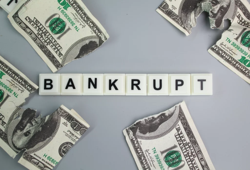 Tiles spelling “BANKRUPT” surrounded by torn 100-dollar bills on a gray background, symbolizing financial ruin and the challenges of multiple bankruptcy filings.