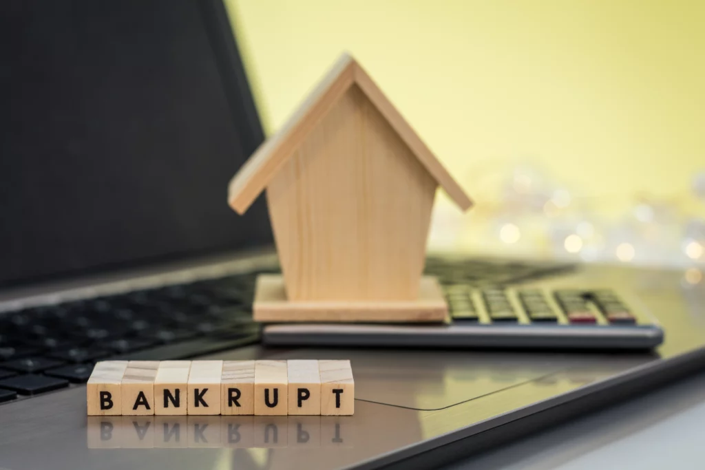 A small wooden house model and calculator on a laptop, with ‘Bankrupt’ spelled out in letter tiles in the foreground.
