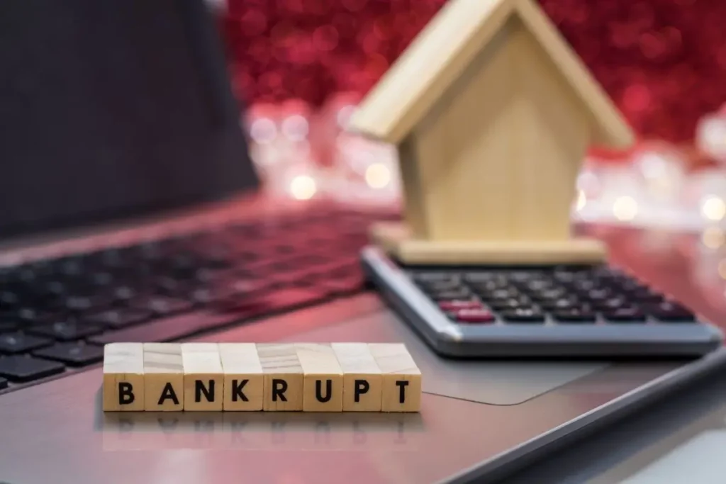 Wooden blocks spelling ‘Bankrupt’ placed on a laptop beside a calculator and wooden house model, symbolizing financial planning and bankruptcy.