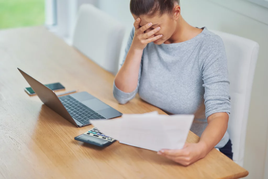 Woman in gray shirt at a wooden table with laptop, calculator, and phone, holding a paper and looking stressed, symbolizing financial hardship.