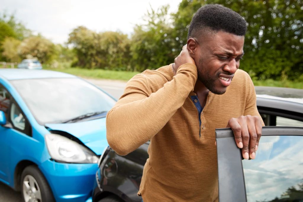 Man in a brown sweater stands by a black car, rubbing his neck in pain after a rear-end collision with a blue car—indicating a recent accident.
