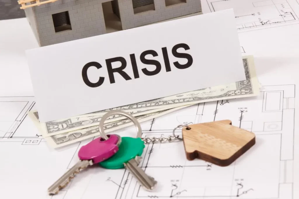 House keys, cash, and a model home beside a sign reading ‘Crisis,’ symbolizing financial trouble and business bankruptcy concerns.